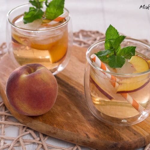 Lillet Rosé White Peach Cocktail 3 Zwei Gläser mit Lillet Rose, Eis und Minze stehen auf einem Holzbrett neben einem frischen Pfirsich.