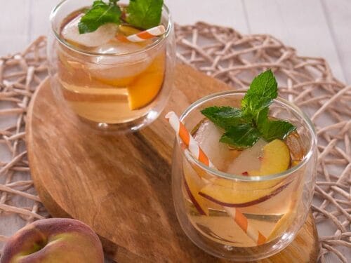 Zwei Gläser mit Lillet Rose White Peach und Minze auf einem Holzbrett, serviert mit frischen Pfirsichspalten.