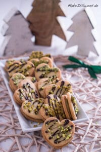 Pistazien Plätzchen mit Mandeln 4 Ein rechteckiger weißer Teller zeigt herzförmige Pistazienplätzchen mit Schokoladenstreifen und gehackten Pistazien, die auf einem Tischset mit hölzernem Baumschmuck im Hintergrund liegen.