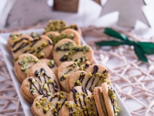 Herzförmige Plätzchen mit Pistazien-Füllung mit Schokoladenüberzug und gehackten Pistazien auf einem weißen Tablett.