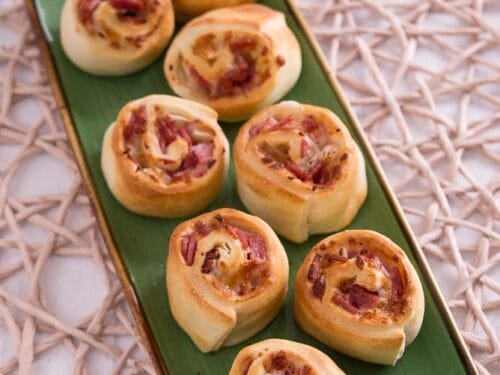 Auf einem rechteckigen grünen Teller liegen acht gebackene Schinken-Käse-Pizzaschnecken aus der Heißluftfritteuse.