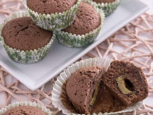 Schokoladenmuffins in Papierförmchen auf einem Teller, eines davon aufgeschnitten, um die gefüllte Pistaziencreme zu zeigen.