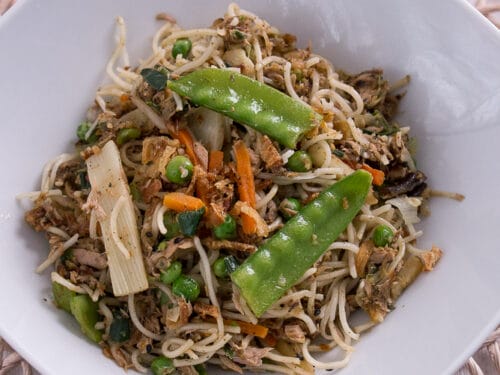 Eine weiße Schale mit Asia-Pfanne, gebratenen Nudeln, Gemüse und grünen Erbsen, die auf einem gewebten Tischset steht.