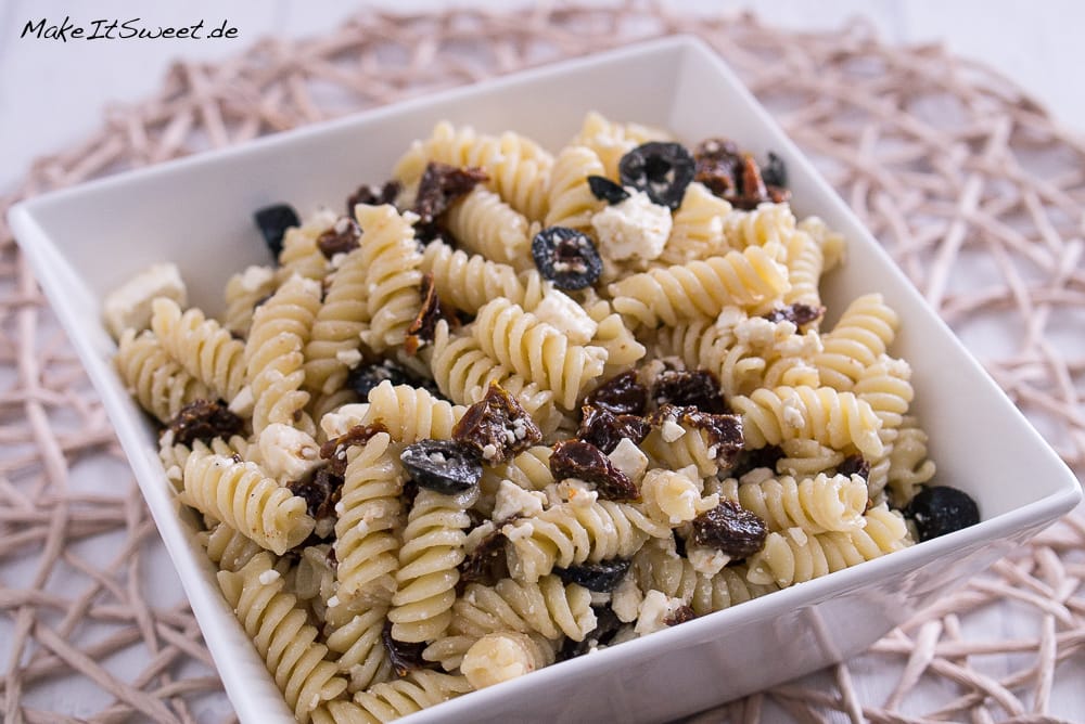 Nudelsalat mit Oliven, sonnengetrockneten Tomaten und Feta-Käse in einer weißen Schale.