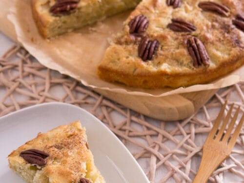 Ein Stück Apfelkuchen mit Pekannüssen auf einem Teller, der restliche Kuchen mit Pekannüssen auf einem Holzbrett dahinter.