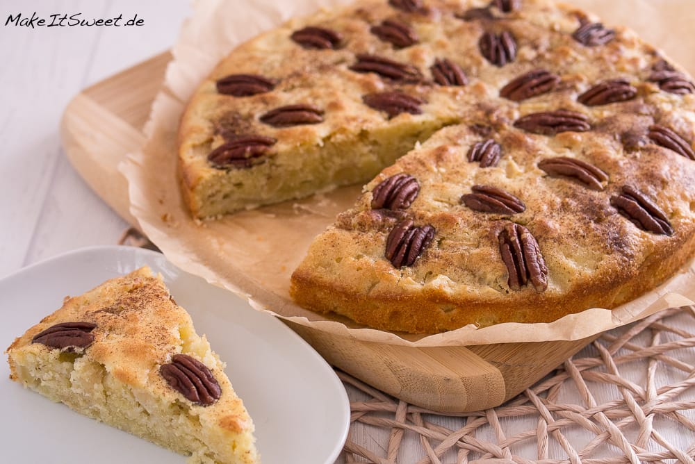 Apfelkuchen mit sichtbaren Pekannüssen auf der Oberseite, eine Scheibe ausgeschnitten, auf Backpapier ausgelegt.