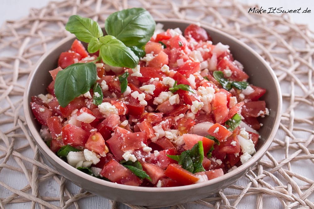 Schale mit frischem Tomatensalat mit Feta, Basilikumblättern und Zwiebeln auf einem gewebten Tischset.