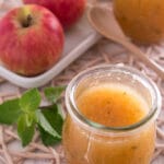 Ein mit Apfelmarmelade gefülltes Glas steht auf einer gewebten Matte. Im Hintergrund sind zwei rote Äpfel auf einer weißen Schale, ein Holzlöffel, ein Minzzweig und ein weiteres Glas mit Marmelade zu sehen.
