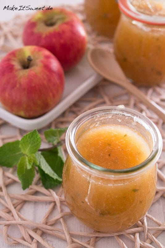 Apfelmarmelade mit Minze 5 Ein mit Apfelmarmelade gefülltes Glas steht auf einer gewebten Matte. Im Hintergrund sind zwei rote Äpfel auf einer weißen Schale, ein Holzlöffel, ein Minzzweig und ein weiteres Glas mit Marmelade zu sehen.