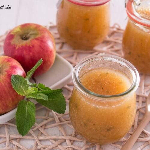 Apfelmarmelade mit Minze 6 Ein Glas mit hellbrauner Apfelmarmelade steht geöffnet mit einem Holzlöffel daneben, daneben zwei rote Äpfel mit grünen Minzblättern und weitere Gläser mit automatisch gespeicherter Entwurfsmarmelade im Hintergrund auf einem gewebten Tischset.