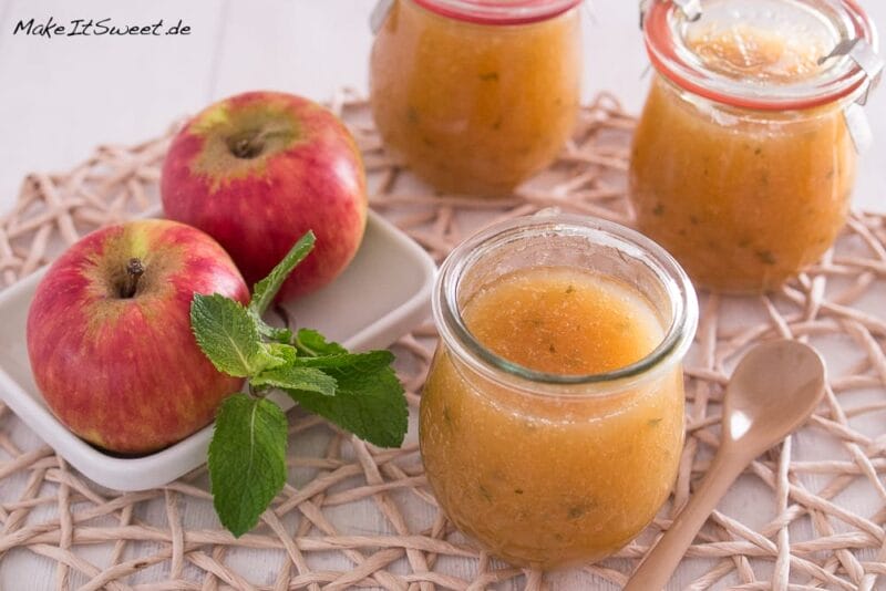 Apfelmarmelade mit Minze 4 Ein Glas mit hellbrauner Apfelmarmelade steht geöffnet mit einem Holzlöffel daneben, daneben zwei rote Äpfel mit grünen Minzblättern und weitere Gläser mit automatisch gespeicherter Entwurfsmarmelade im Hintergrund auf einem gewebten Tischset.