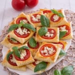 Ein Teller mit quadratischen Blätterteig-Törtchen, belegt mit Tomatenscheiben, zerbröseltem Feta und frischen Basilikumblättern, steht auf einer gewebten Matte. Zwei ganze Tomaten und zusätzliche Basilikumblätter sind daneben angeordnet.