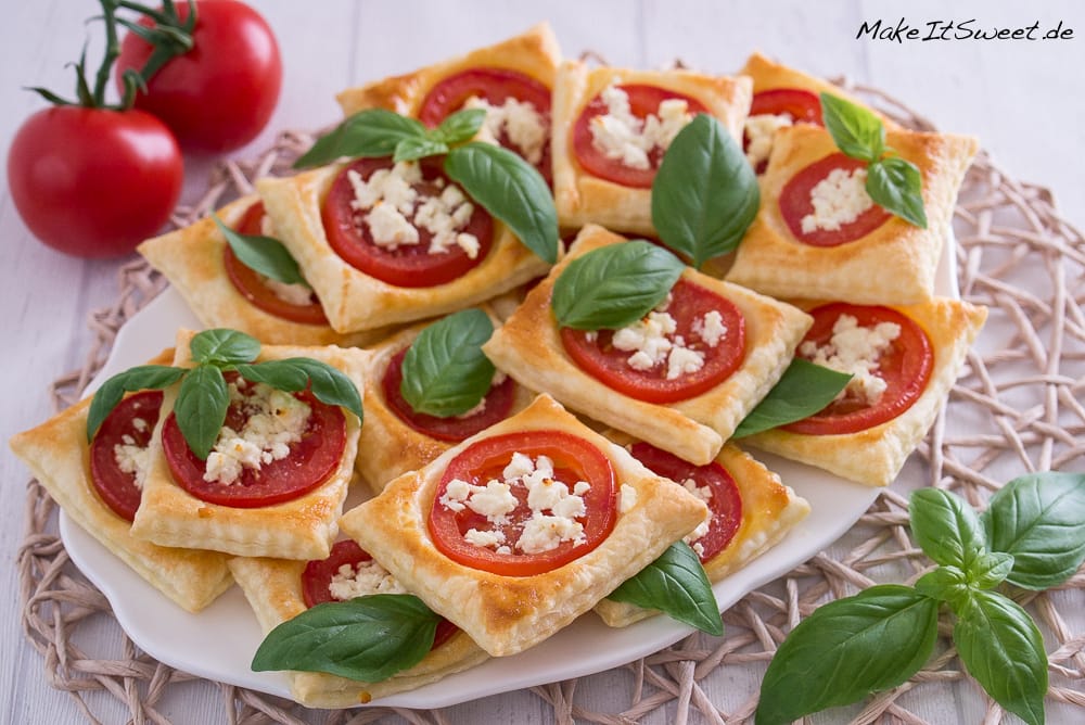 Quadratischer Blätterteig mit Tomatenscheiben, zerbröckeltem Käse und Basilikumblättern auf einem weißen Teller.