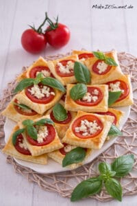 Ein Teller mit quadratischen Blätterteig-Törtchen, belegt mit Tomatenscheiben, zerbröseltem Feta und frischen Basilikumblättern, steht auf einer gewebten Matte. Zwei ganze Tomaten und zusätzliche Basilikumblätter sind daneben angeordnet.
