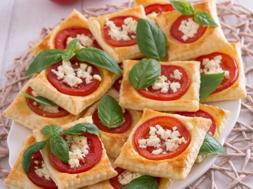 Quadratische Blätterteig-Teigtaschen mit Tomate, Feta und Basilikum auf weißem Teller.