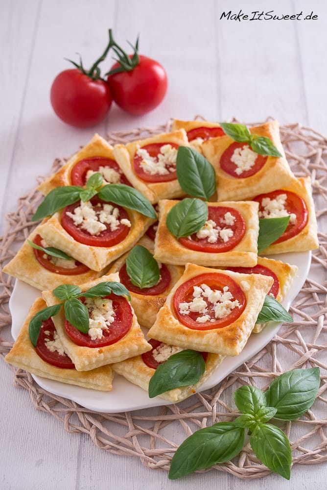 Ein Teller mit quadratischen Blätterteig-Törtchen, belegt mit Tomatenscheiben, zerbröseltem Feta und frischen Basilikumblättern, steht auf einer gewebten Matte. Zwei ganze Tomaten und zusätzliche Basilikumblätter sind daneben angeordnet.