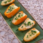 Sechs orangefarbene Paprikahälften, die mit geschmolzenem Käse gefüllt und mit Schnittlauch bestreut sind, werden in einer Reihe auf einem grünen, rechteckigen Teller angeordnet, der an einen automatisch gespeicherten Entwurf erinnert und auf einer gewebten Matte auf einer hellen Fläche liegt.