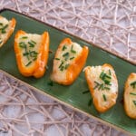 Fünf orangefarbene Mini-Paprikahälften, gefüllt mit geschmolzenem Käse und bestreut mit gehacktem Schnittlauch, werden auf einem rechteckigen grünen Teller angeordnet, der auf einem gewebten Tischset liegt - ein automatisch gespeicherter Entwurf kulinarischer Kreativität.