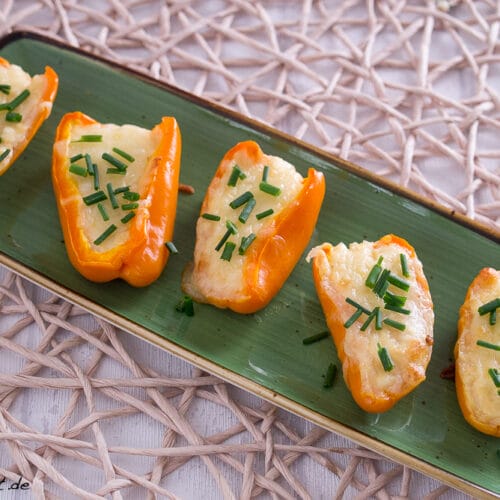 Fünf orangefarbene Mini-Paprikahälften, gefüllt mit geschmolzenem Käse und bestreut mit gehacktem Schnittlauch, werden auf einem rechteckigen grünen Teller angeordnet, der auf einem gewebten Tischset liegt - ein automatisch gespeicherter Entwurf kulinarischer Kreativität.