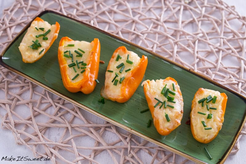 Fünf orangefarbene Mini-Paprikahälften, gefüllt mit geschmolzenem Käse und bestreut mit gehacktem Schnittlauch, werden auf einem rechteckigen grünen Teller angeordnet, der auf einem gewebten Tischset liegt - ein automatisch gespeicherter Entwurf kulinarischer Kreativität.