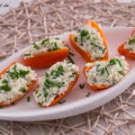 Sechs orangefarbene Paprikahälften, gefüllt mit einer weißen Käse-Kräuter-Mischung, garniert mit Schnittlauch, werden auf einem weißen ovalen Teller auf einem gewebten Tischset angerichtet - ein automatisch gespeicherter Entwurf für mühelose Unterhaltung.