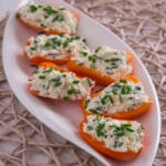 Mini-Paprika gefüllt mit Feta und Schnittlauch Sechs halbierte orangefarbene Mini-Paprikaschoten sind mit einer Hüttenkäse-Schnittlauch-Mischung gefüllt und auf einem weißen ovalen Teller auf einer gewebten Matte fein säuberlich angeordnet - ein automatisch gespeicherter Entwurf frischer Aromen und einer lebendigen Präsentation.