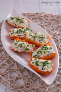 Mini-Paprika gefüllt mit Feta und Schnittlauch 6 Sechs halbierte orangefarbene Mini-Paprikaschoten sind mit einer Hüttenkäse-Schnittlauch-Mischung gefüllt und auf einem weißen ovalen Teller auf einer gewebten Matte fein säuberlich angeordnet - ein automatisch gespeicherter Entwurf frischer Aromen und einer lebendigen Präsentation.