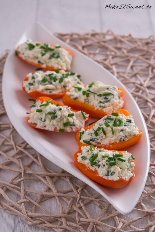 Mini-Paprika gefüllt mit Feta und Schnittlauch 2 Sechs halbierte orangefarbene Mini-Paprikaschoten sind mit einer Hüttenkäse-Schnittlauch-Mischung gefüllt und auf einem weißen ovalen Teller auf einer gewebten Matte fein säuberlich angeordnet - ein automatisch gespeicherter Entwurf frischer Aromen und einer lebendigen Präsentation.
