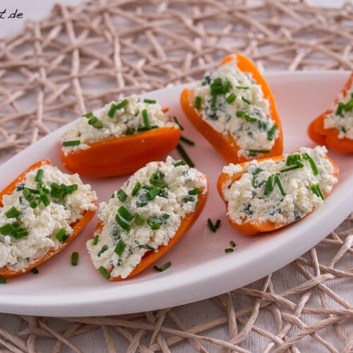 Mini-Paprika gefüllt mit Feta und Schnittlauch 3 Sechs orangefarbene Paprikahälften, gefüllt mit einer weißen Käse-Kräuter-Mischung, garniert mit Schnittlauch, werden auf einem weißen ovalen Teller auf einem gewebten Tischset angerichtet - ein automatisch gespeicherter Entwurf für mühelose Unterhaltung.
