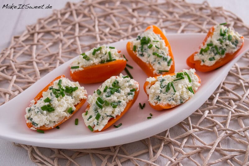 Mini-Paprika gefüllt mit Feta und Schnittlauch 1 Sechs orangefarbene Paprikahälften, gefüllt mit einer weißen Käse-Kräuter-Mischung, garniert mit Schnittlauch, werden auf einem weißen ovalen Teller auf einem gewebten Tischset angerichtet - ein automatisch gespeicherter Entwurf für mühelose Unterhaltung.