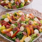 Zwei Teller mit Salat, Mozzarellakugeln, Nektarinenscheiben und Prosciutto sind auf einem Tischset angeordnet. Die Salate werden in weißen Schalen serviert und mit zusätzlichen Salatblättern garniert.