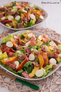 Zwei Teller mit Salat, Mozzarellakugeln, Nektarinenscheiben und Prosciutto sind auf einem Tischset angeordnet. Die Salate werden in weißen Schalen serviert und mit zusätzlichen Salatblättern garniert.