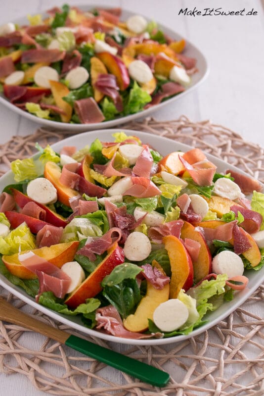 Nektarinen-Mozzarella-Salat mit Serrano Schinken 2 Zwei Teller mit Salat, Mozzarellakugeln, Nektarinenscheiben und Prosciutto sind auf einem Tischset angeordnet. Die Salate werden in weißen Schalen serviert und mit zusätzlichen Salatblättern garniert.