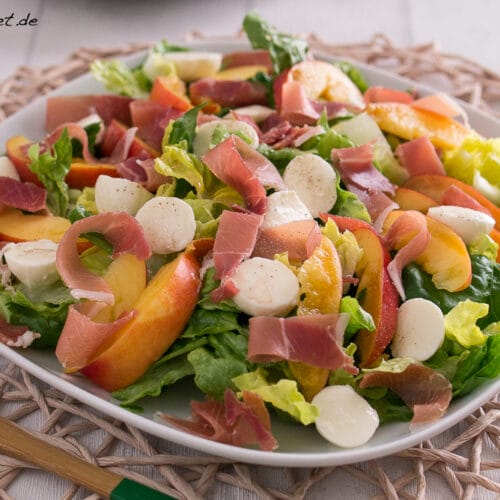 Nektarinen-Mozzarella-Salat mit Serrano Schinken 3 Ein Teller bunter Salat mit frischen Nektarinen, Mozzarella-Bällchen und feinem Aufschnitt auf einem geflochtenen Platzset, daneben ein Holzgabel. "MakeItSweet.de" steht oben links.