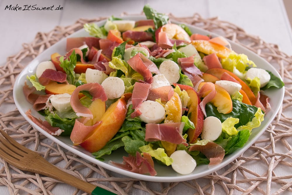 Nektarinen-Mozzarella-Salat mit Kopfsalat, Pfirsichscheiben, Mozzarellakugeln und Prosciutto auf einem weißen Teller.