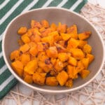 Eine beigefarbene Schale mit gebratenen Kürbis- und Süßkartoffelwürfeln steht auf einem Tischset aus Weidengeflecht, daneben liegt ein grün-weiß gestreiftes Handtuch auf einer weißen Holzfläche. Perfekt für jedes Kürbisrezept mit Ihrer Heißluftfritteuse.