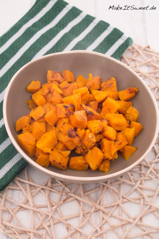 Kürbis aus der Heißluftfritteuse 2 Eine beigefarbene Schale mit gebratenen Kürbiswürfeln steht auf einem Tischset, daneben liegt ein grün-weiß gestreiftes Handtuch auf einer weißen Holzfläche. Perfekt für jedes Kürbisrezept mit Ihrer Heißluftfritteuse.