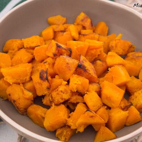 Kürbis aus der Heißluftfritteuse 3 Eine Schale mit gerösteten, gewürfelten Orangenkürbissen steht auf einem geflochtenen Tischset neben einem grün-weiß gestreiften Tuch - ein automatisch gespeicherter Entwurf von gemütlichen Herbstaromen.