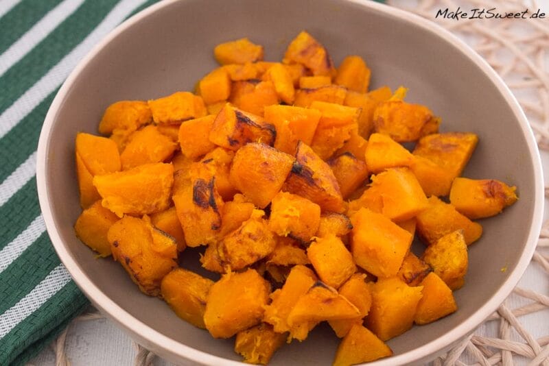 Kürbis aus der Heißluftfritteuse 1 Eine Schale mit gerösteten, gewürfelten Kürbissen steht auf einem geflochtenen Tischset neben einem grün-weiß gestreiften Tuch - ein automatisch gespeicherter Entwurf von gemütlichen Herbstaromen.