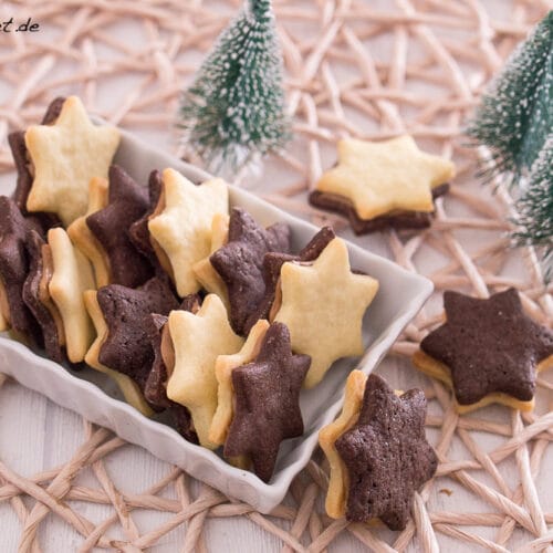 Eine rechteckige Schale enthält sternförmige Plätzchen mit Schokoladen- und Vanilleschichten und einer cremigen Füllung, die auf einer gewebten Matte stehen. Im Hintergrund stehen drei kleine künstliche Tannenbäume.