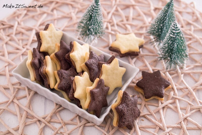 Eine rechteckige Schale enthält sternförmige Plätzchen mit Schokoladen- und Vanilleschichten und einer cremigen Füllung, die auf einer gewebten Matte stehen. Im Hintergrund stehen drei kleine Tannenbäume.