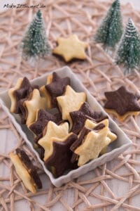 Plätzchen mit Erdnussbutter Füllung 6 Eine rechteckige Schale, gefüllt mit sternförmigen Keksen, einige aus Natur und einige aus Schokolade, steht auf einem gewebten Tischset neben kleinen dekorativen Tannenbäumen.