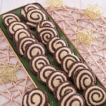 Auf einem rechteckigen grünen Teller liegen Reihen von Marmorkeksen mit Schokoladen- und Vanillekipferln. Der Teller steht auf einem gewebten Tischset mit goldenen Sternen, das auf einer hellen Holzoberfläche steht.