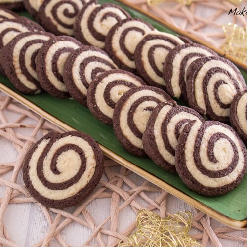 Auf einem rechteckigen grünen Teller liegt eine Reihe von Schwarz-Weiß-Gebäck, ein klassisches deutsches Gebäck mit Schokoladen- und Vanillekipferln, fein säuberlich angeordnet. Der Teller steht auf einem geflochtenen Tischset mit dekorativen goldenen Sternen in der Nähe.