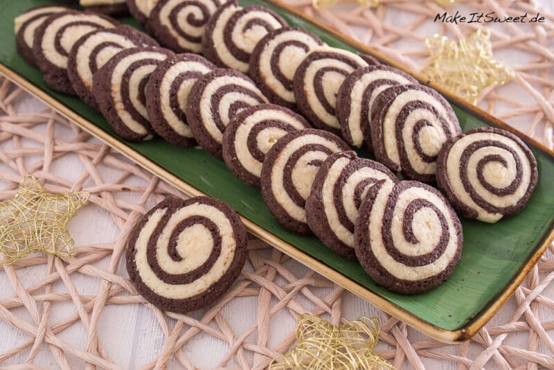 Auf einem rechteckigen grünen Teller liegt eine Reihe von Schwarz-Weiß-Gebäck, fein säuberlich angeordnet. Der Teller steht auf einem geflochtenen Tischset mit dekorativen goldenen Sternen in der Nähe.