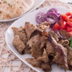 Ein Teller mit in Scheiben geschnittenem Dönerfleisch, gehackten roten Zwiebeln, Tomaten und Gurken liegt neben rundem Brot auf einem separaten Teller, alles auf einem gewebten Tischset angeordnet - ein automatisch gespeicherter Entwurf einer köstlichen Mahlzeit.