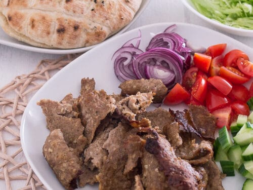 Teller mit Dönerfleisch, geschnittenen roten Zwiebeln, Tomaten, Gurken, Salat und Fladenbrot im Hintergrund.