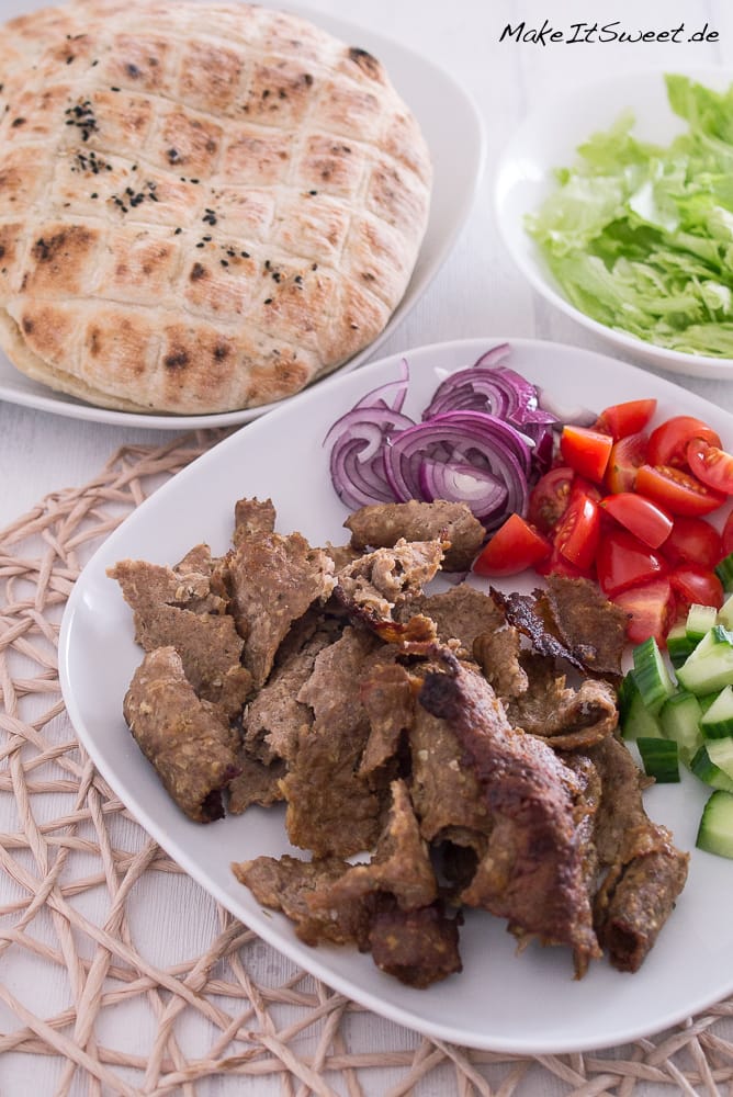 Backpapier Döner aus der Heißluftfriteusse 1 Ein Teller mit in Scheiben geschnittenem gekochtem Dönerfleisch, gehackten roten Zwiebeln, gewürfelten Tomaten, Gurkenstücken und geschreddertem Salat, daneben ein rundes Fladenbrot mit schwarzen Kernen, alles auf einem gewebten Platzdeckchen. Perfekt zum Genießen nach dem Knuspern in der Heißluftfriteuse.