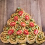 Ein weihnachtlicher Pesto-Tannenbaum aus herzhaften Pinwheel-Rollen auf einem hölzernen Schneidebrett, dekoriert mit halbierten Kirschtomaten und gehackten grünen Zwiebeln - perfektes Fingerfood Weihnachten für Ihren Festtagstisch.