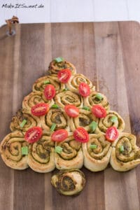 Ein weihnachtlicher Pesto-Tannenbaum aus herzhaften Pinwheel-Rollen auf einem hölzernen Schneidebrett, dekoriert mit halbierten Kirschtomaten und gehackten grünen Zwiebeln - perfektes Fingerfood Weihnachten für Ihren Festtagstisch.